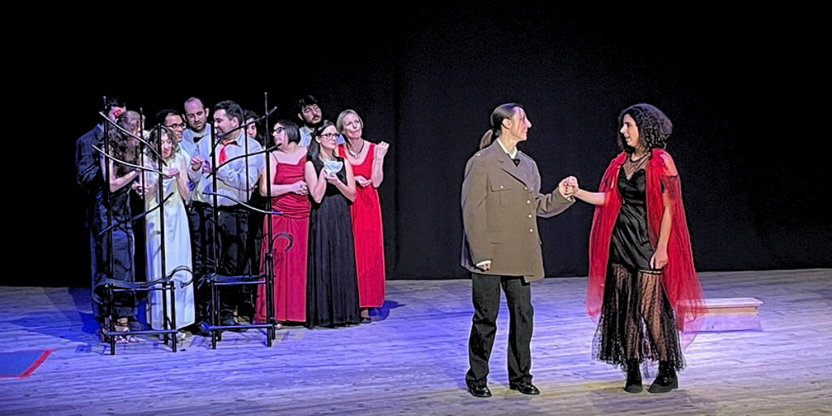 Foto di una scena dello spettacolo Triangoli del gruppo studentesco Teatro a Rotelle a Teatro Camploy, con due ragazze che si tengono per mano e un gruppo di attori dietro ad un oggetto di scena.