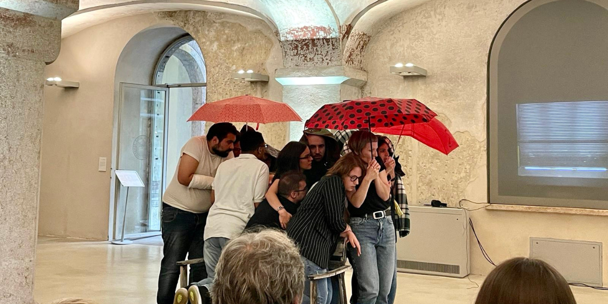 Snaphot of actors taking shelter under red umbrellas in the production of In principio era la Rupe